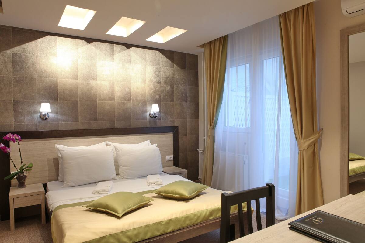 A hotel room with a bed, bedside lamps, a nightstand, a chair, a table, and a window with curtains.