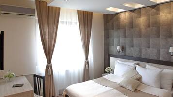 A modern hotel room with a large bed, a desk, and a chair. There is an air conditioning unit on the wall, and the room has a neutral color scheme.