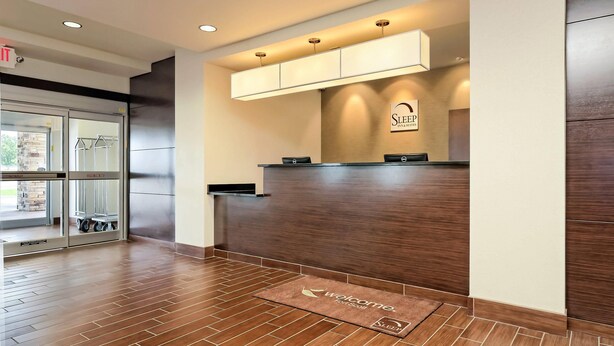 A reception area with a wooden reception desk, a glass door entrance, and a welcome mat. - Sleep Inn & Suites Fort Scott (Fort Scott)