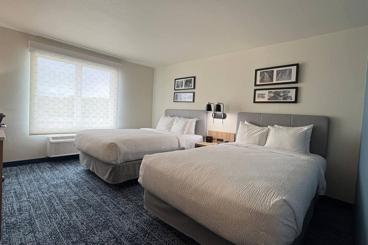 A hotel room with two beds, a window with blinds, a bedside table with a lamp, and framed pictures on the wall.