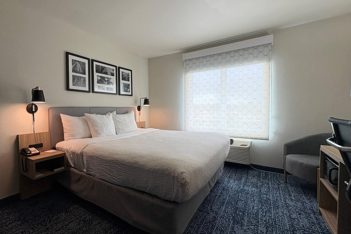 A hotel room with a bed, bedside tables, a desk, a chair, a TV, and a window with patterned curtains.