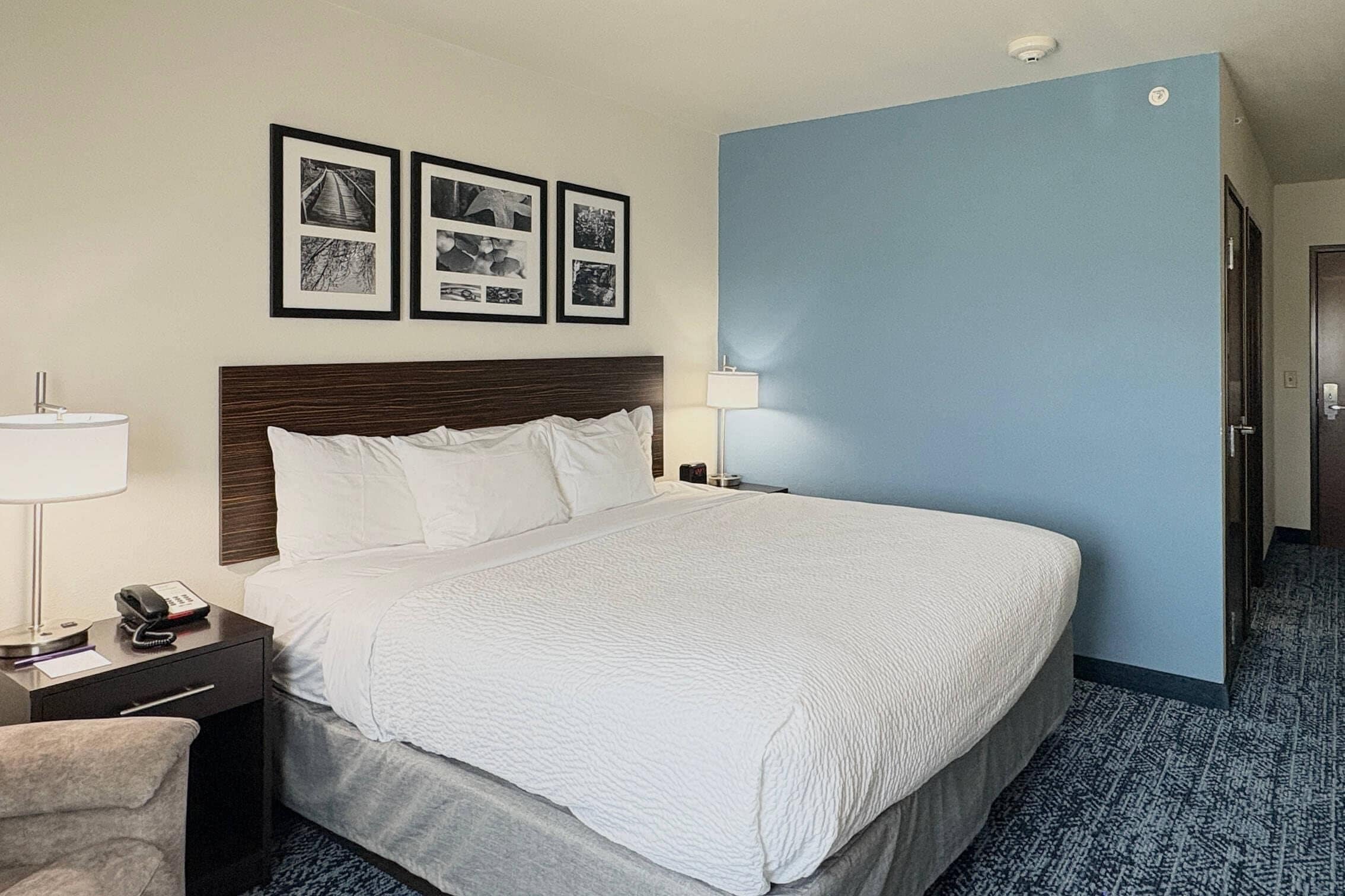 A hotel room with a large bed, a nightstand with a lamp and phone, a chair, and framed pictures on the wall.