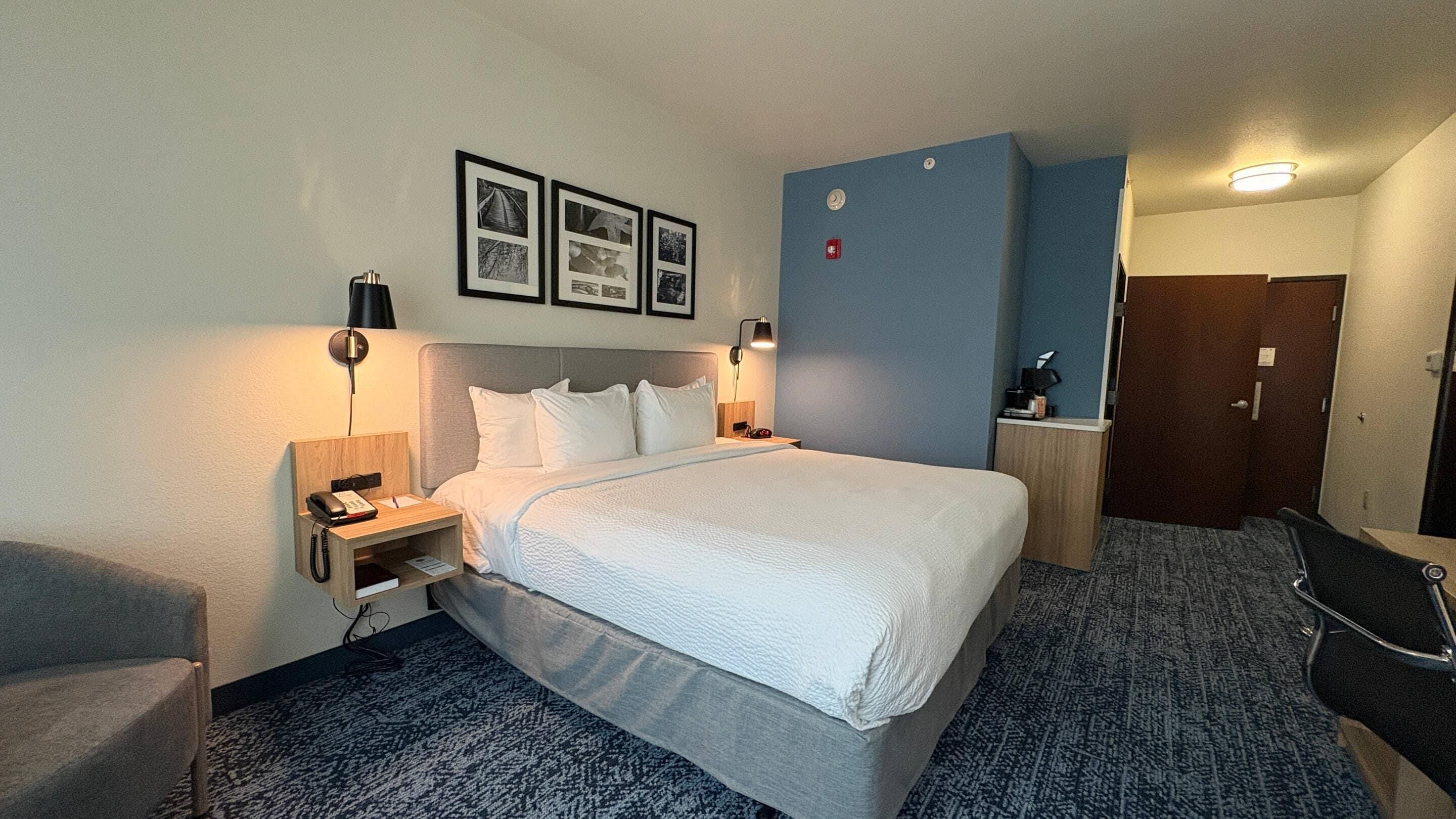A hotel room with a bed, bedside tables, a desk, a chair, a wall-mounted phone, and framed pictures.