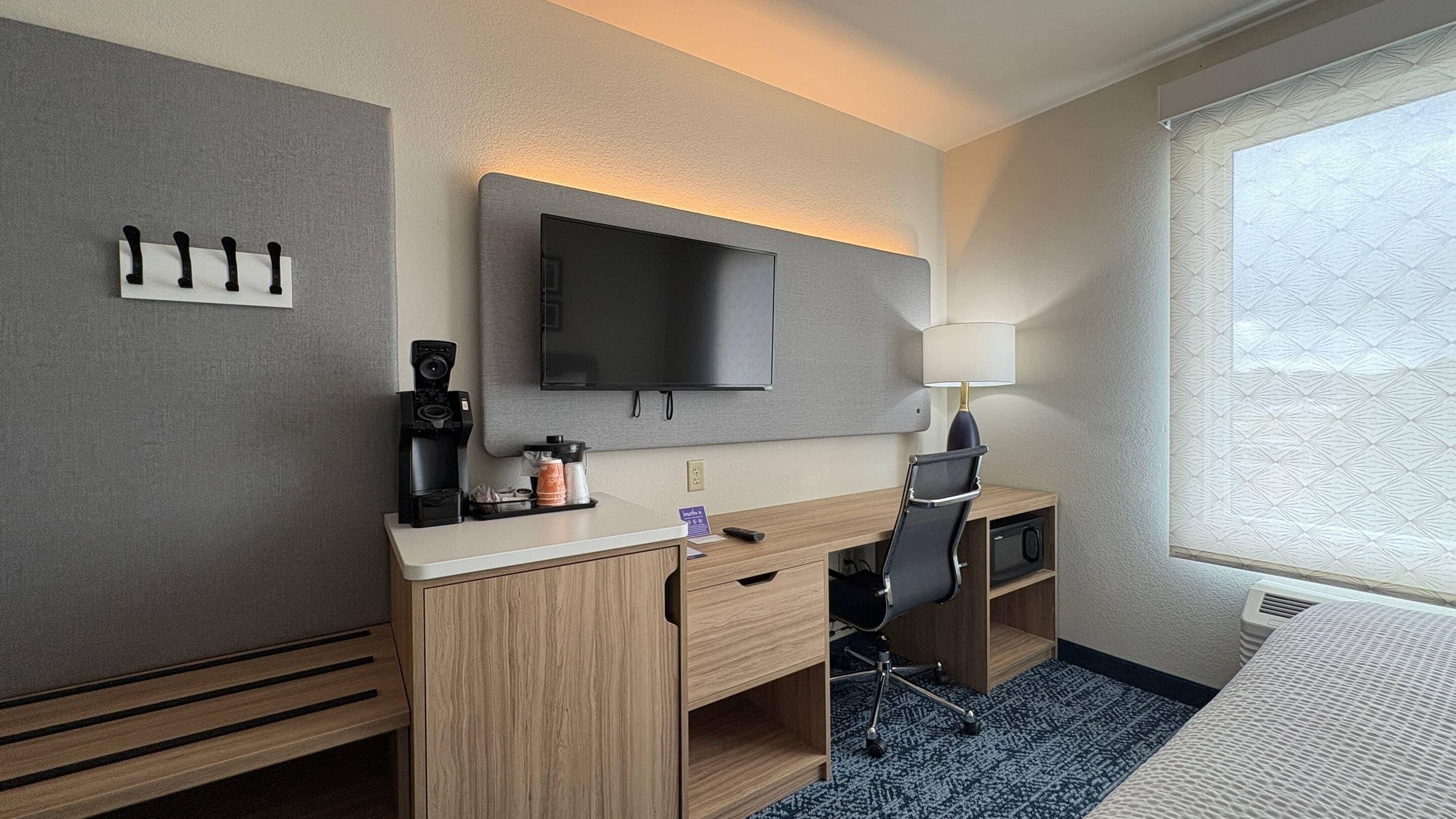 A hotel room with a flat-screen TV, a desk with a coffee maker, a lamp, and a bed.