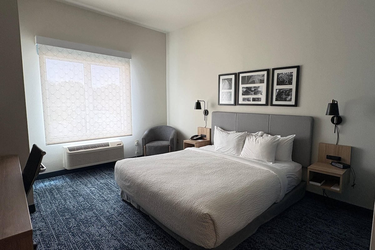 A hotel room with a large bed, a chair, a desk, a window with blinds, and framed pictures on the wall.