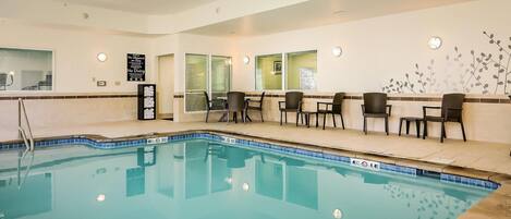 An indoor swimming pool with a tiled edge, surrounded by lounge chairs and a wall with a clock and a sign.