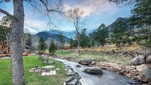 A river flowing through a landscaped area with a wooden deck, outdoor seating, and a mountain backdrop. - 4 Seasons Inn on Fall River (Estes Park)