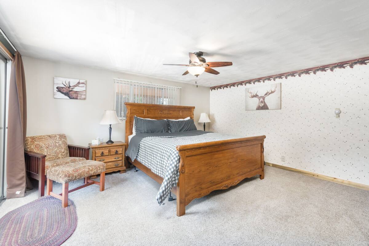A bedroom with a wooden bed, a chair, a nightstand, a ceiling fan, and a window with blinds.