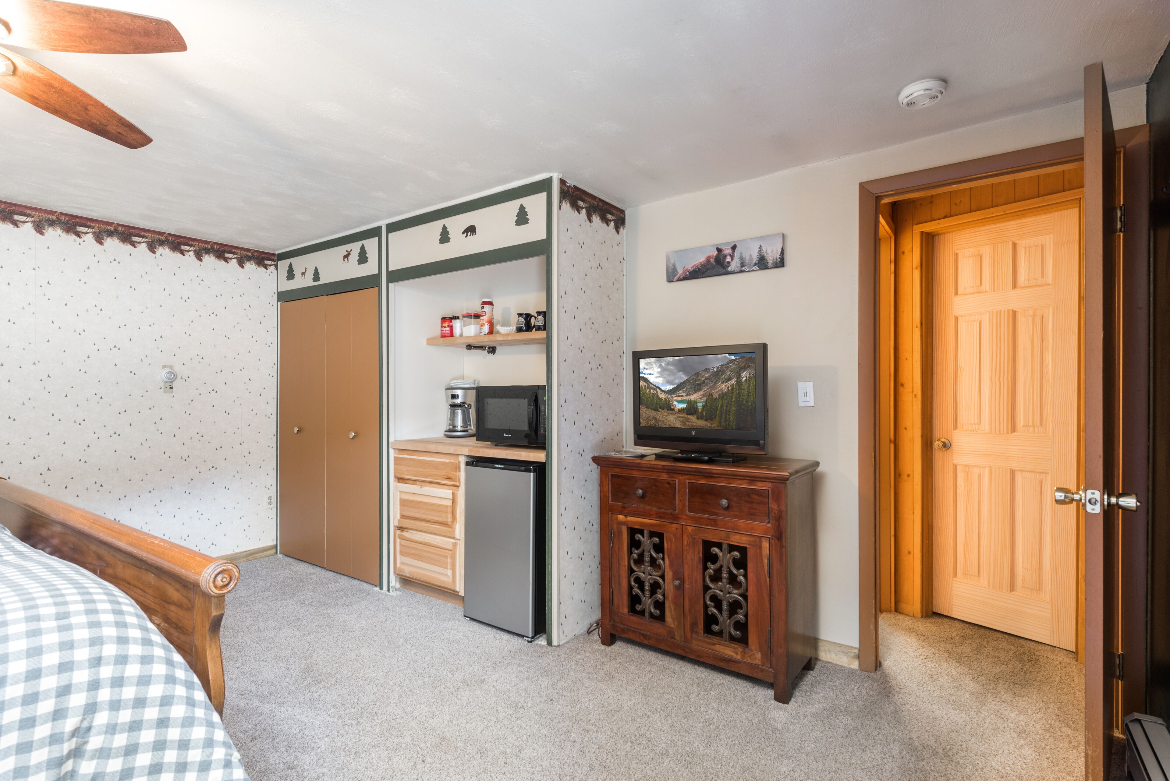 A bedroom with a bed, a wooden wardrobe, a small kitchenette with a microwave, a refrigerator, and a television on a wooden cabinet.