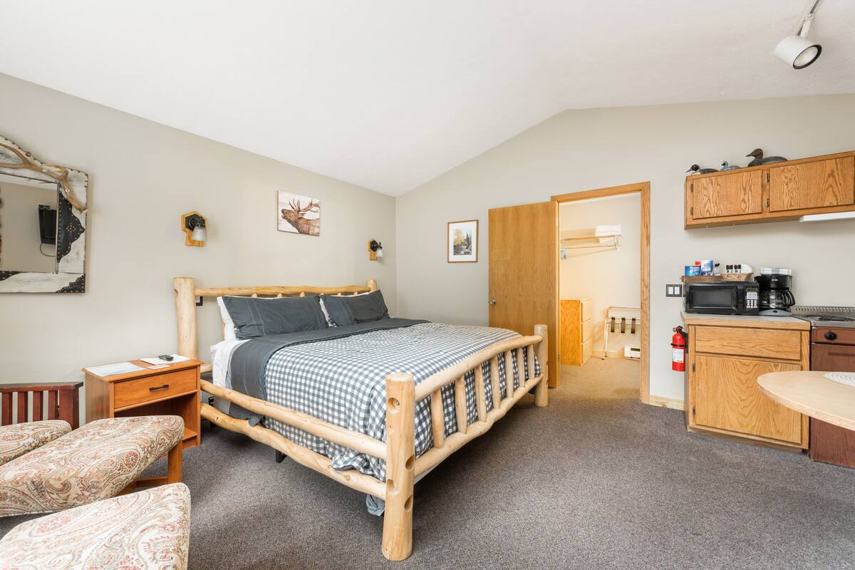A bedroom with a wooden bed, bedside tables, a small desk, a chair, a wardrobe, and a kitchenette with a microwave and coffee maker.