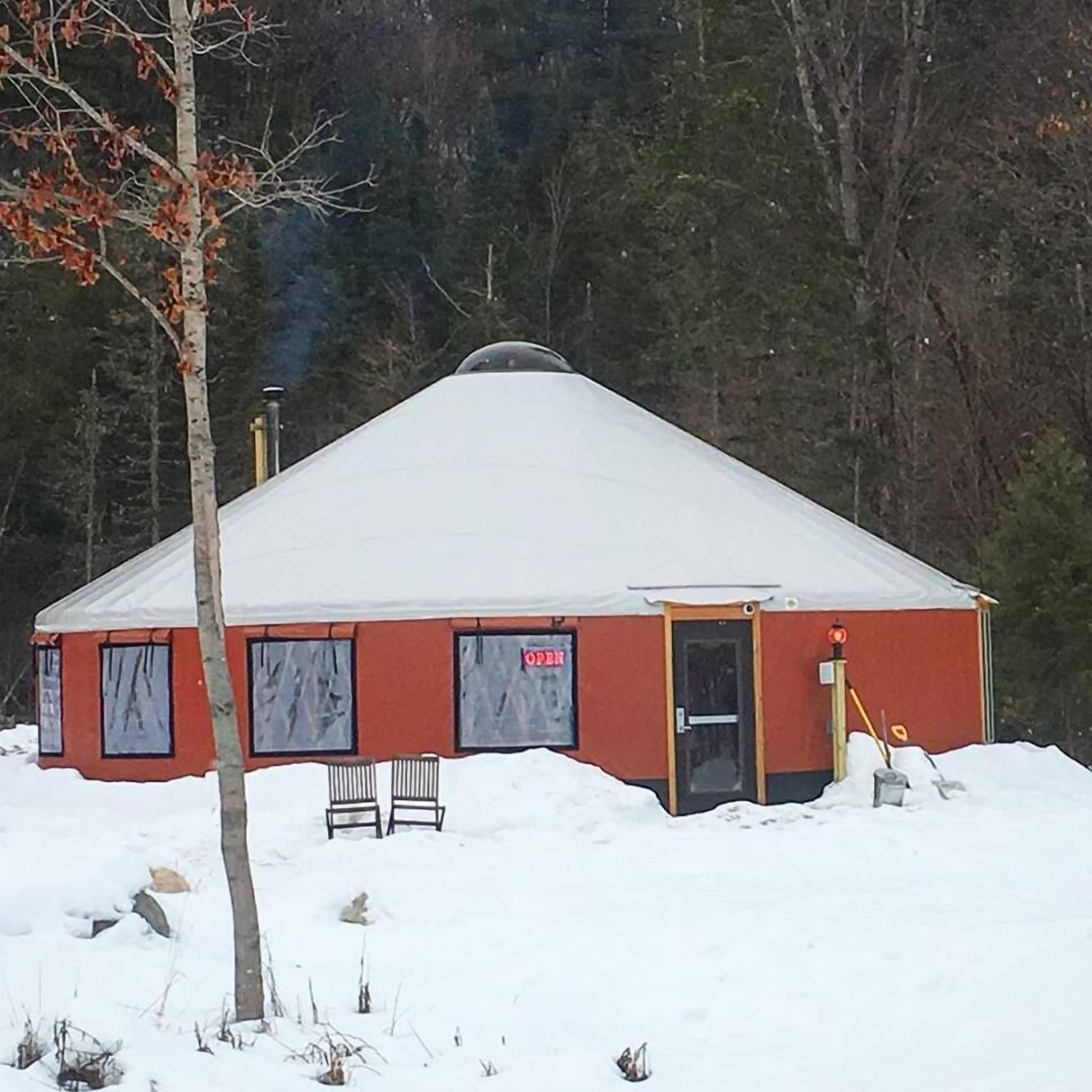 Una yurta cubierta de nieve con exterior rojo y techo blanco, ubicada en una zona boscosa.