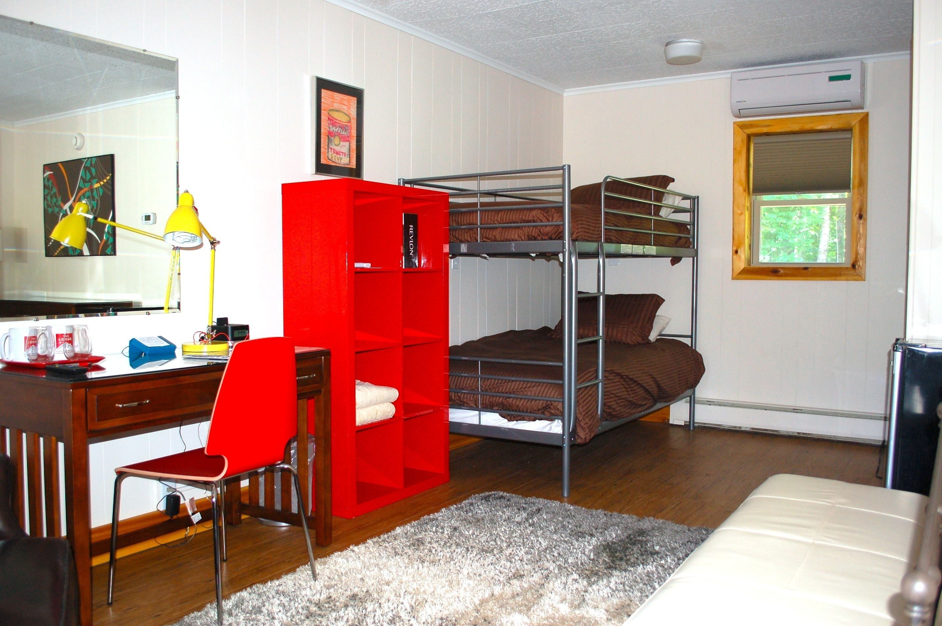Un dormitorio con literas, un escritorio con silla, un estante rojo, un espejo y una ventana.