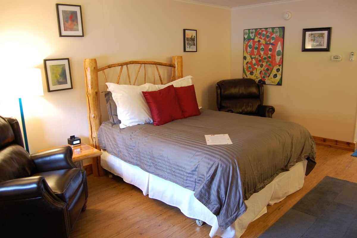 A bedroom with a wooden headboard, a bed with gray and white bedding, a red pillow, a brown leather chair, and a small table with a clock.