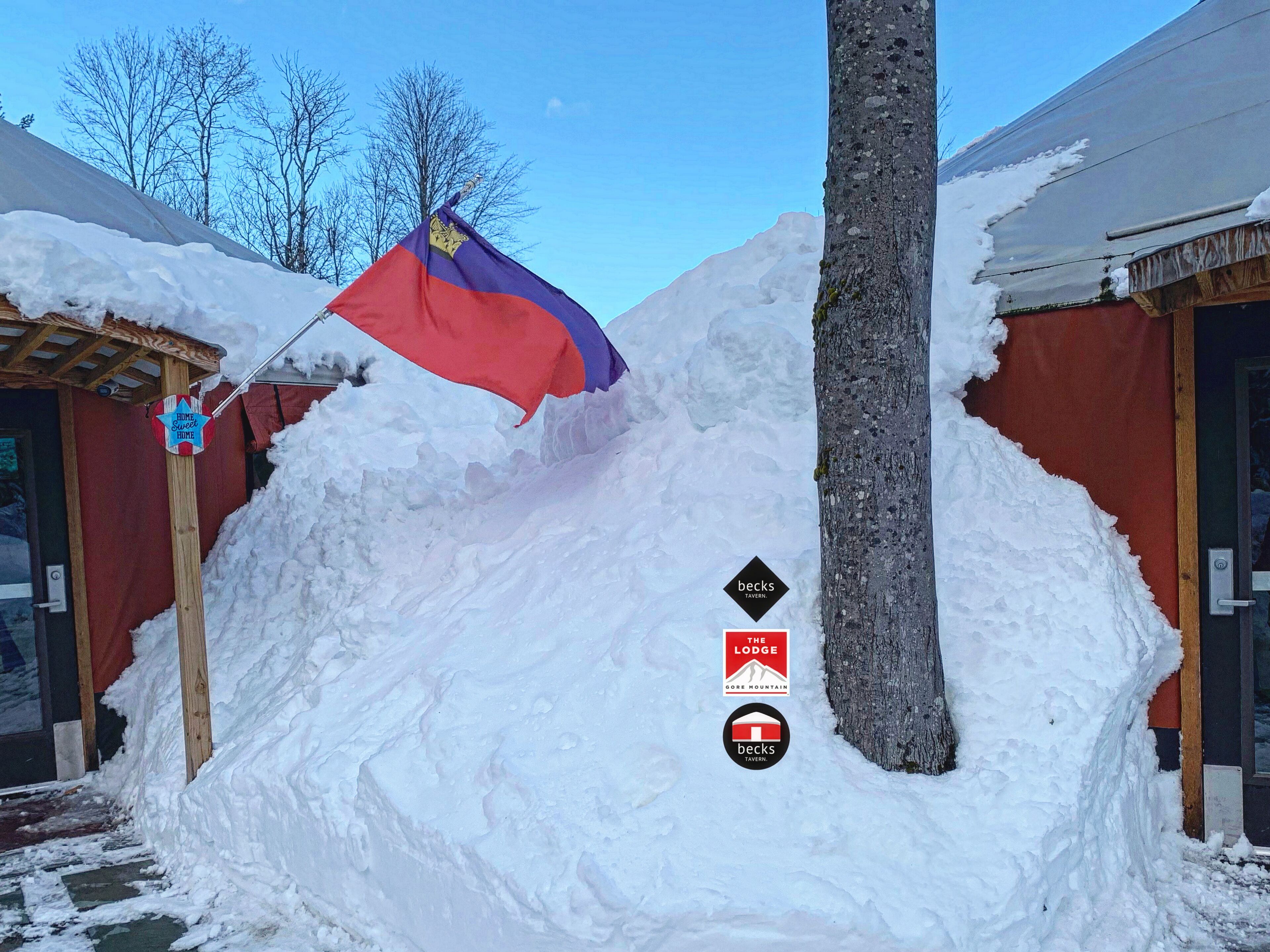 Zona exterior coberta de neu amb una bandera, indicadors per a 'Home Switch Inn' i 'Becks Tavern', i un arbre.