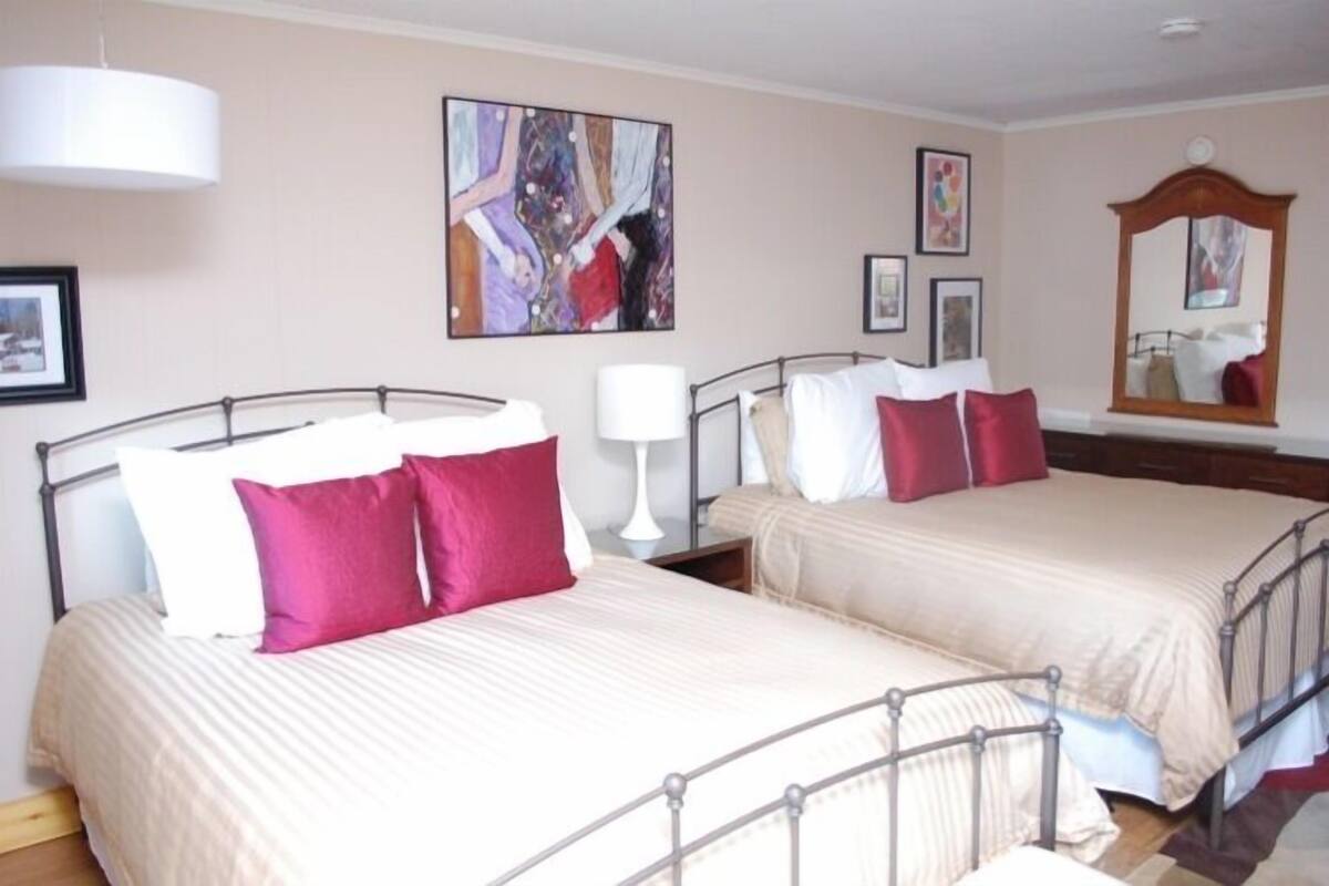 A hotel room with two beds, each with a metal frame and beige bedding. There is a large abstract painting on the wall, a mirror, and a lamp on the nightstand.
