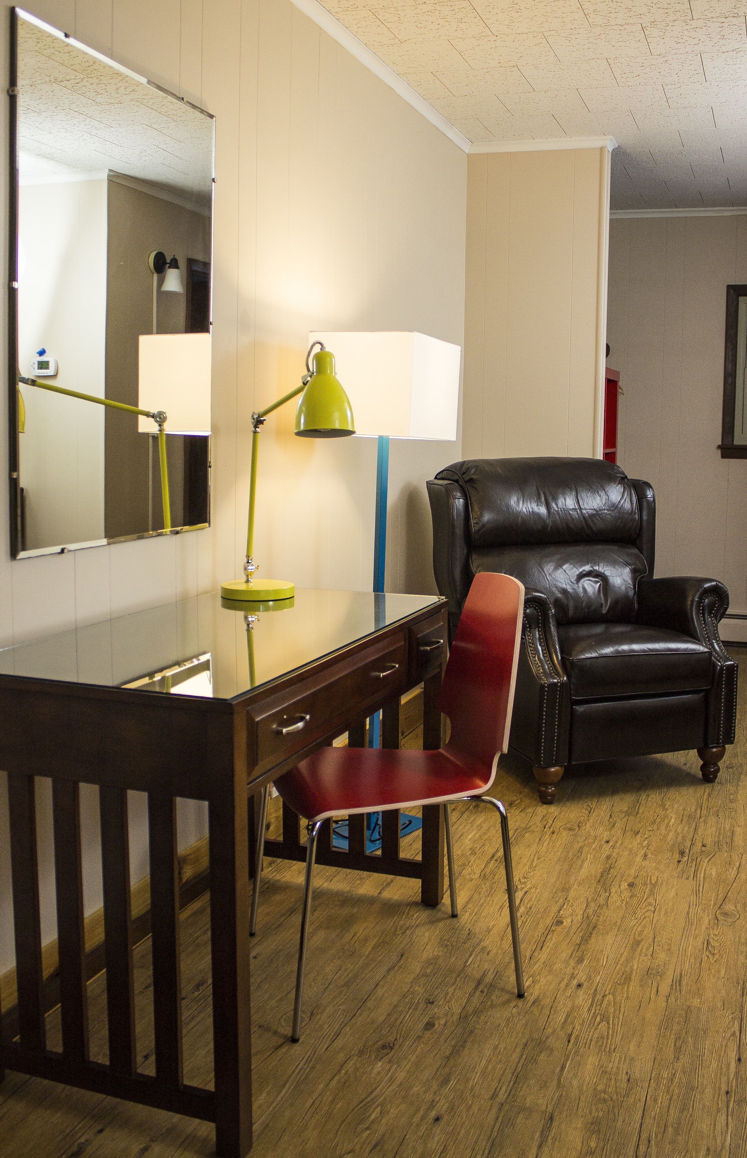 Une pièce comprenant un bureau en bois, une chaise rouge, un fauteuil en cuir noir, un miroir et une lampe de bureau verte.