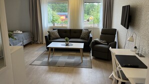Una sala moderna con sofá, sillón y una mesa con lámpara. Hay ventanas con cortinas, un televisor montado en la pared y vistas al exterior con vegetación.