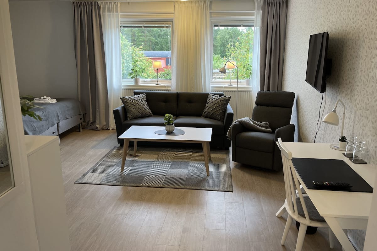 A modern living room with a sofa, armchair, and a table with a lamp. There are windows with curtains, a TV mounted on the wall, and a view of greenery outside.