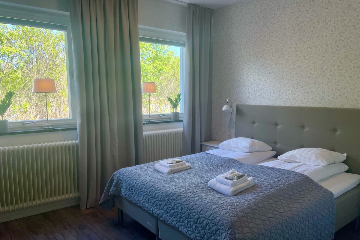 A hotel room with a bed, a window with curtains, a nightstand, a lamp, and a radiator.
