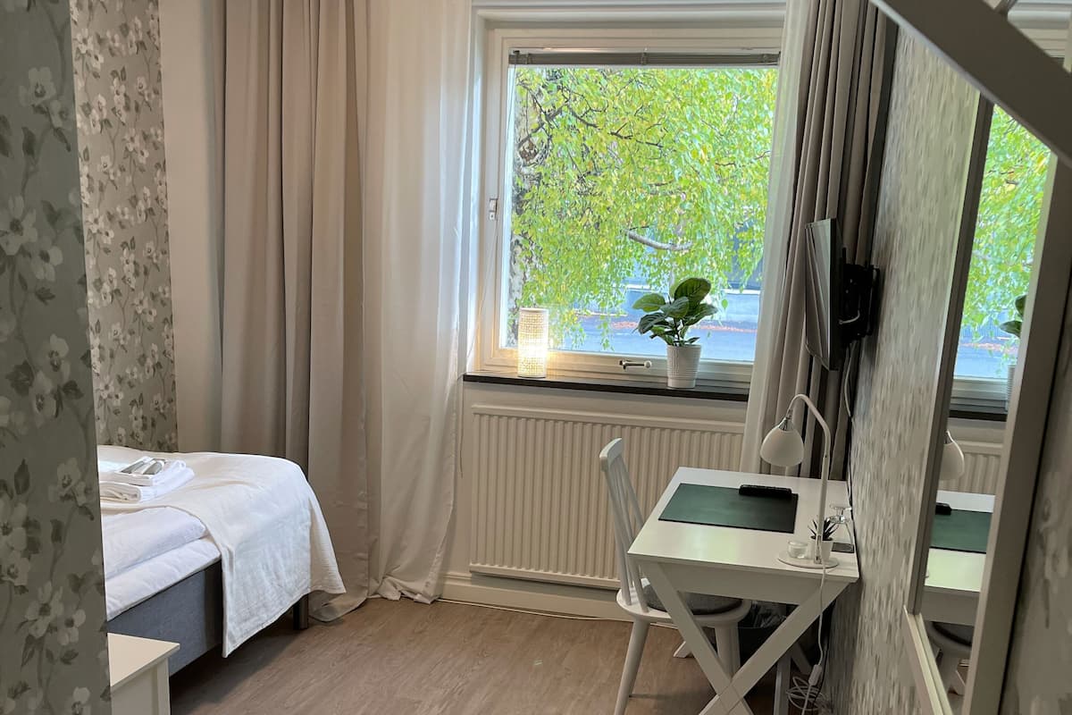 A bedroom with a bed, a desk with a chair, a window with a view of greenery, and a radiator.