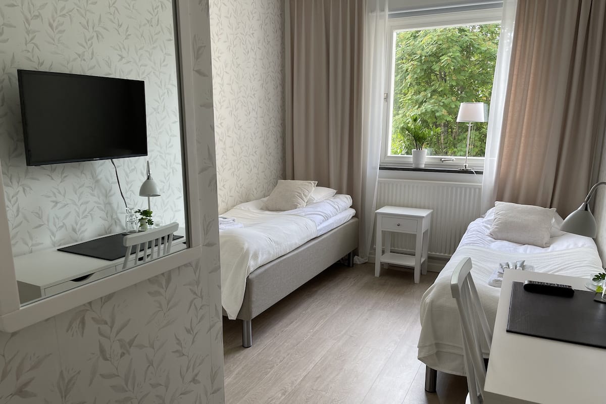 A hotel room with two beds, a desk, a TV, and a window with a view of greenery.