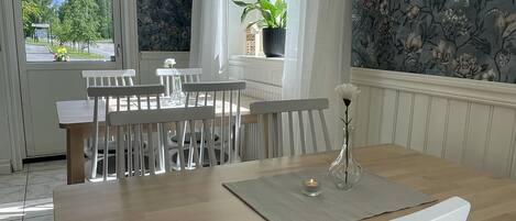 A dining area with a wooden table and white chairs, a potted plant on the windowsill, and a view of the outside.