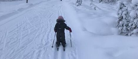 A snowy path with a child skiing, surrounded by snow-covered trees and streetlights.