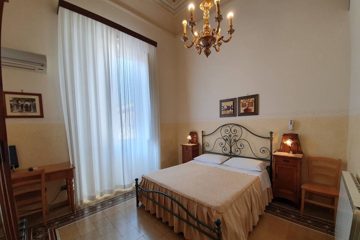 A bedroom with a bed, a chandelier, a nightstand, a chair, and a desk.