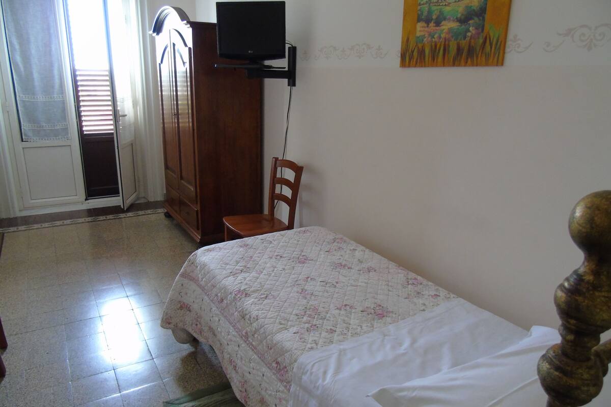 A bedroom with a bed, a wooden wardrobe, a television on a stand, and a chair.