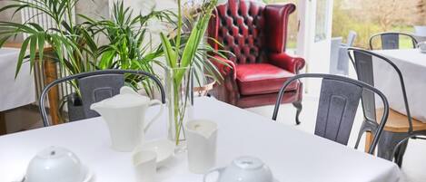 Un coin repas avec une table dressée pour le thé, entourée de chaises, d’un fauteuil rouge à revêtement en tissu et de plantes en pot.