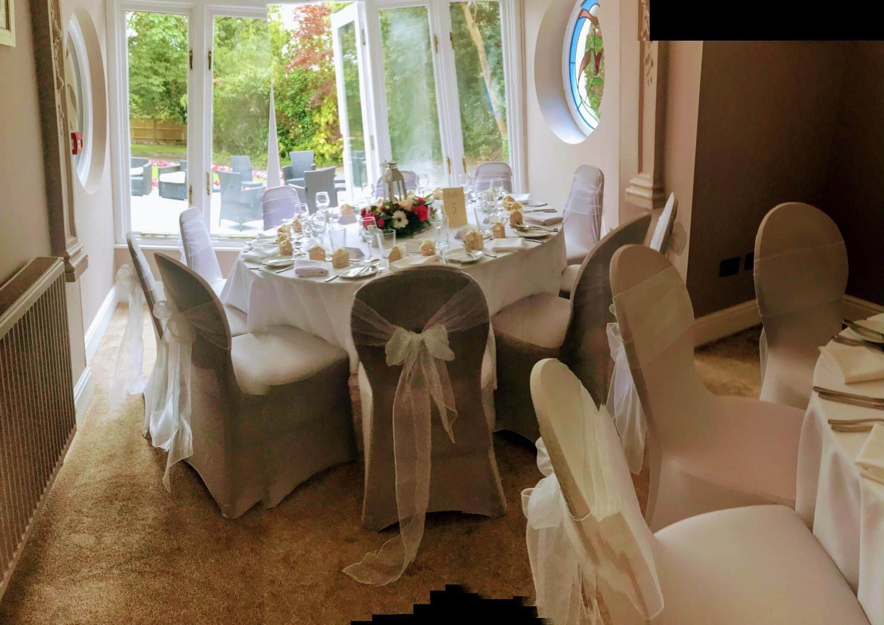A dining area with round tables set for a meal, chairs with white covers, and a view of greenery outside.