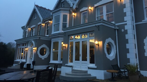 A large, two-story building with a grey facade and white trim, featuring multiple windows and a central entrance with a set of stairs. - Llwyn Country House (Llanelli)