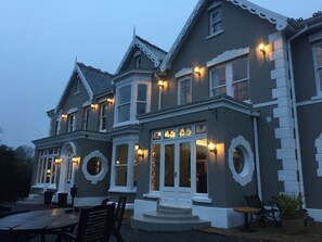 A large, two-story building with a grey facade and white trim, featuring multiple windows and a central entrance with a set of stairs. - Llwyn Country House (Llanelli)