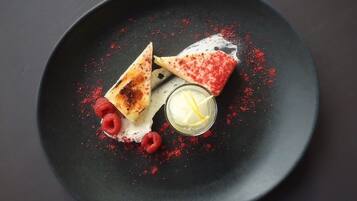 Um prato de sobremesa com um prato escuro, dois doces triangulares, framboesas e um copo pequeno com uma fatia amarela.
