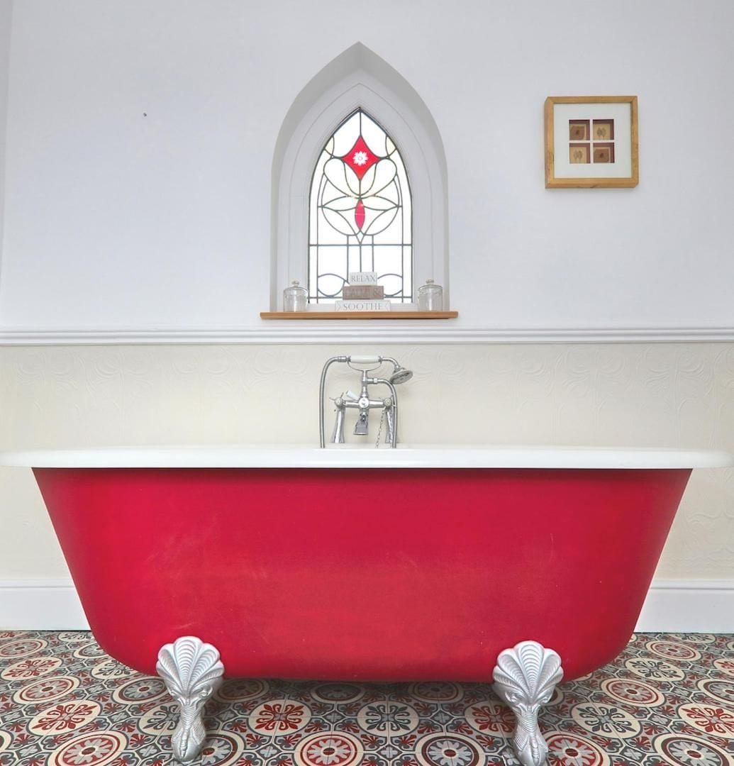 A red clawfoot bathtub in a bathroom with a decorative window and a framed picture on the wall.