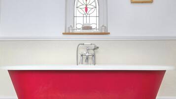 Een rode pootbad in een badkamer met een decoratief raam en een ingelijst schilderij aan de muur.