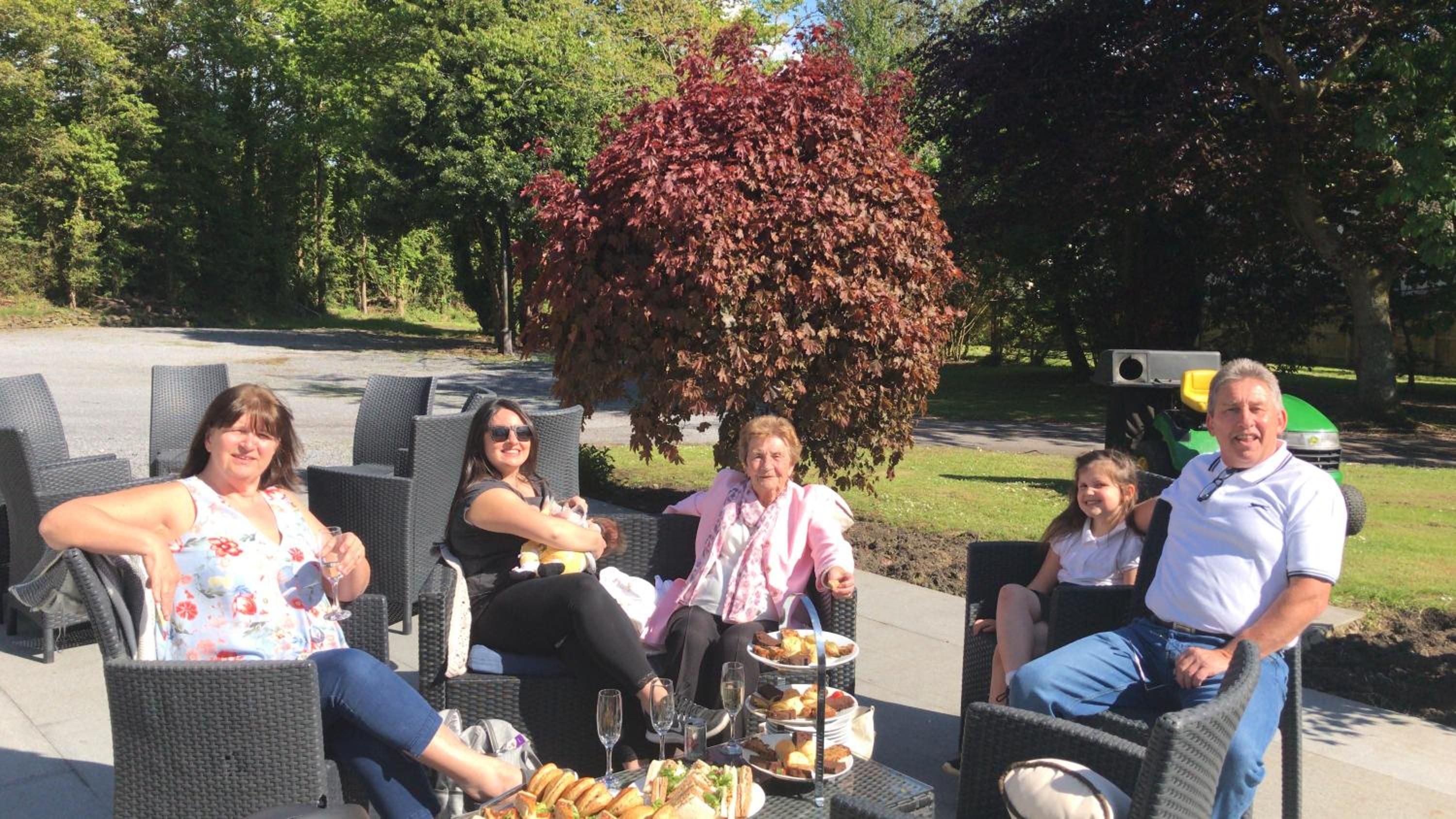 A group of people seated on outdoor wicker chairs with a table of food and drinks in front of them.