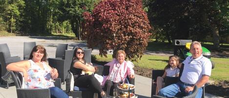 A group of people seated on outdoor wicker chairs with a table of food and drinks in front of them.