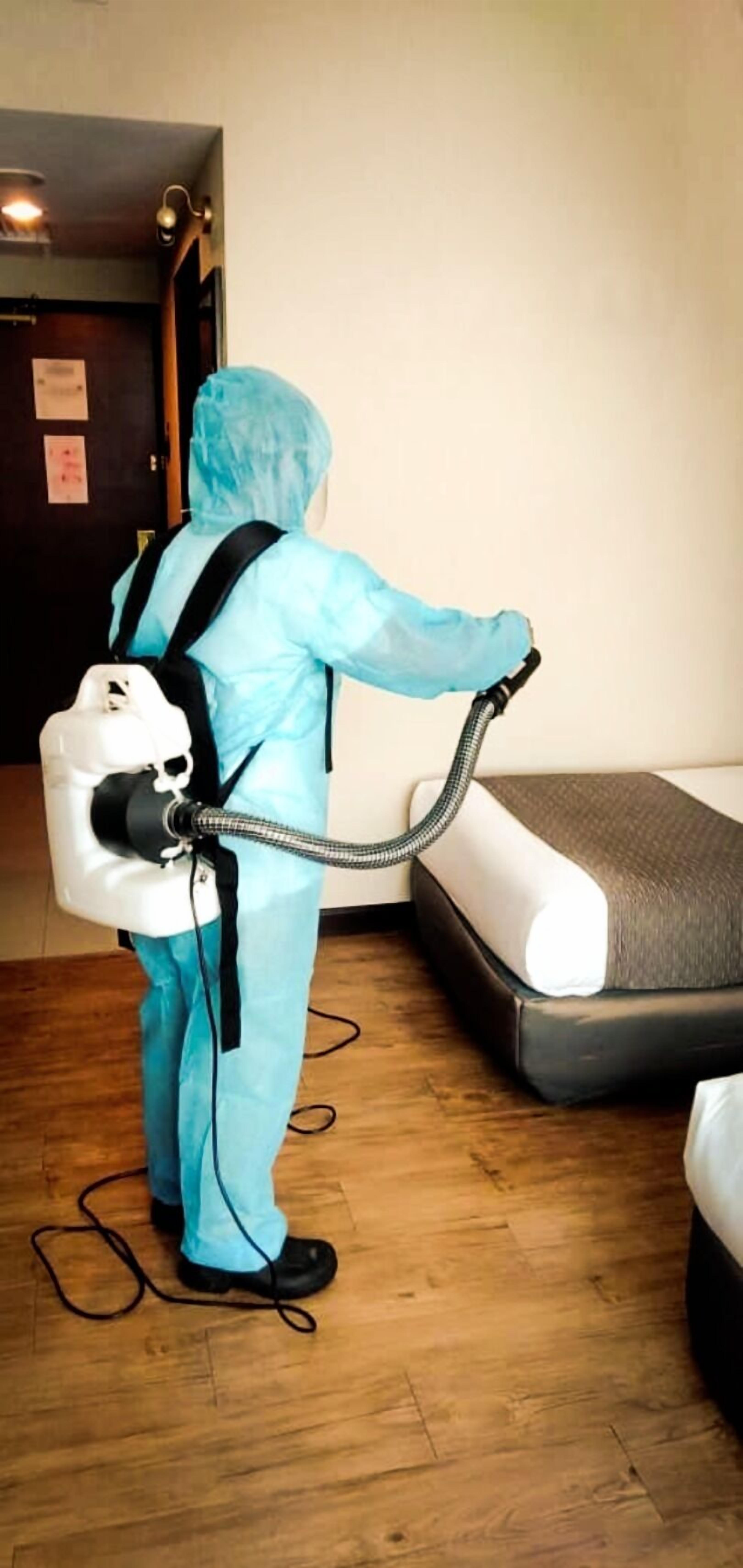 A person in a blue protective suit using a vacuum cleaner on a mattress in a room with wooden flooring and a door in the background.