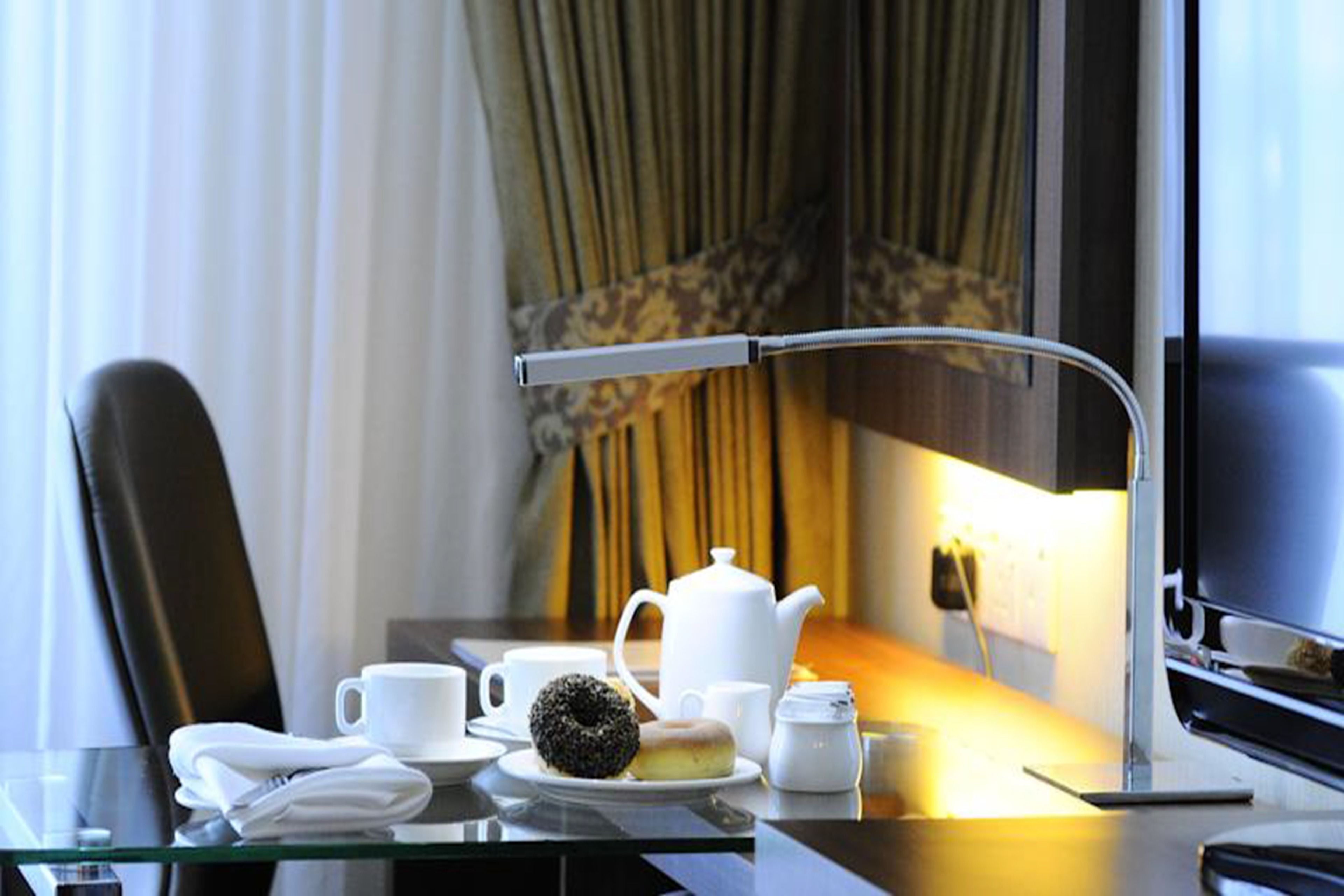 A hotel room with a glass table set for breakfast, including a teapot, cups, and a donut.