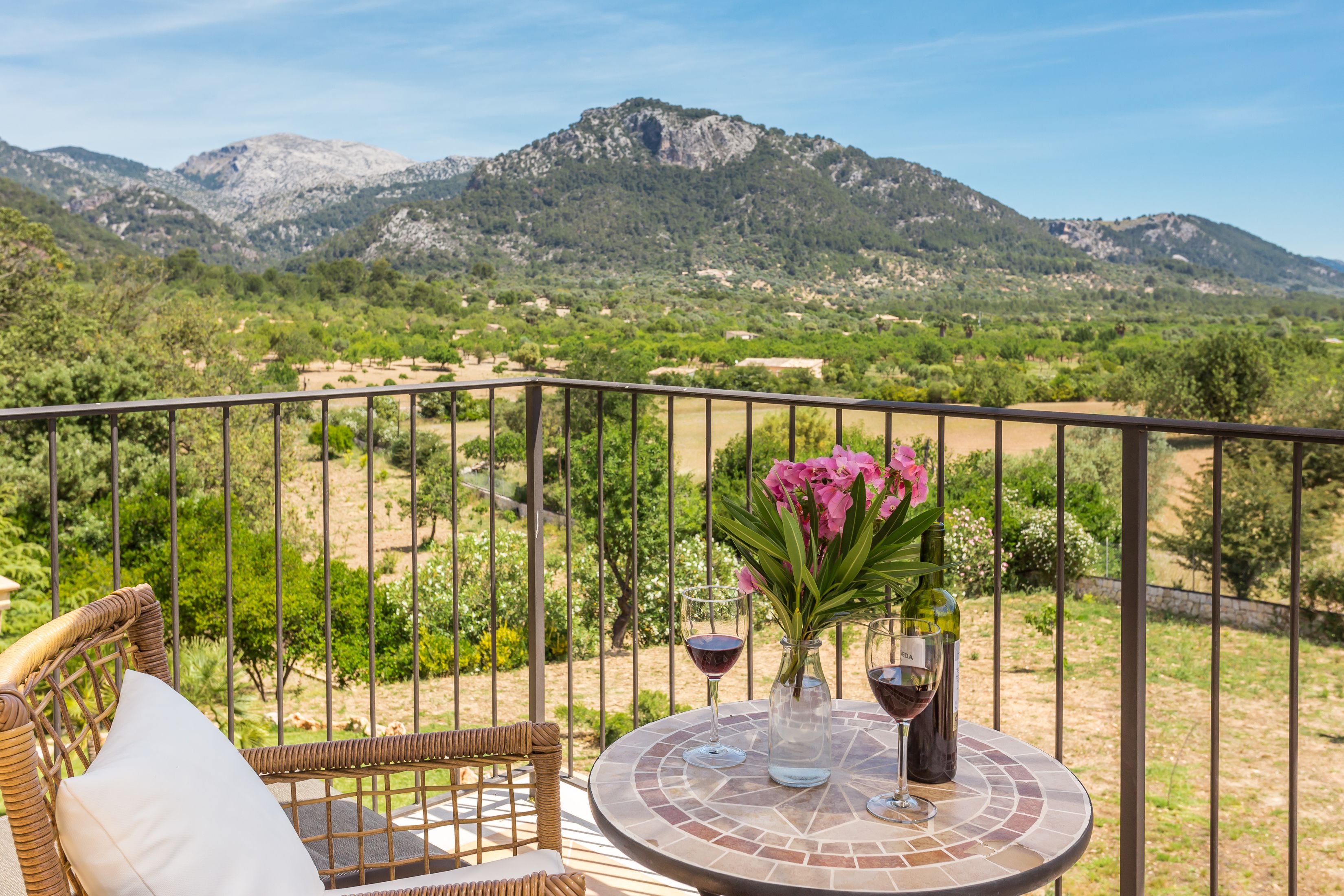 Un balcón con muebles de mimbre y una mesa con copas de vino y un florero, con vista a un paisaje montañoso.