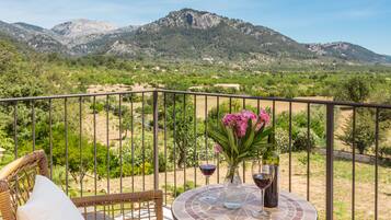 Un balcón con muebles de mimbre y una mesa con copas de vino y un florero, con vista a un paisaje montañoso.