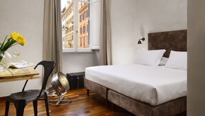 A hotel room with a bed, a table with a chair, a window with curtains, and a view of buildings outside. - San Pietro Boutique Rooms (Rome)