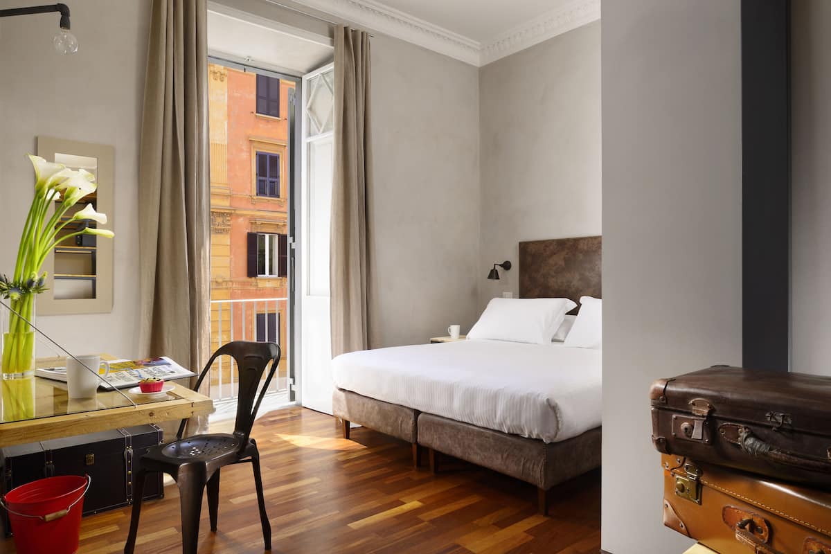 Une chambre d’hôtel avec un lit, un bureau et une chaise, un vase avec des fleurs et une valise.