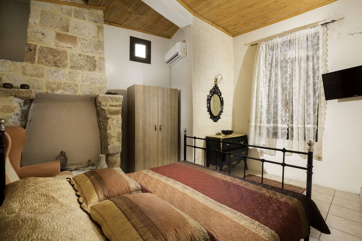 Un dormitorio con pared de piedra, una cama con ropa de cama estampada, un armario de madera, una mesita y un televisor en la pared.