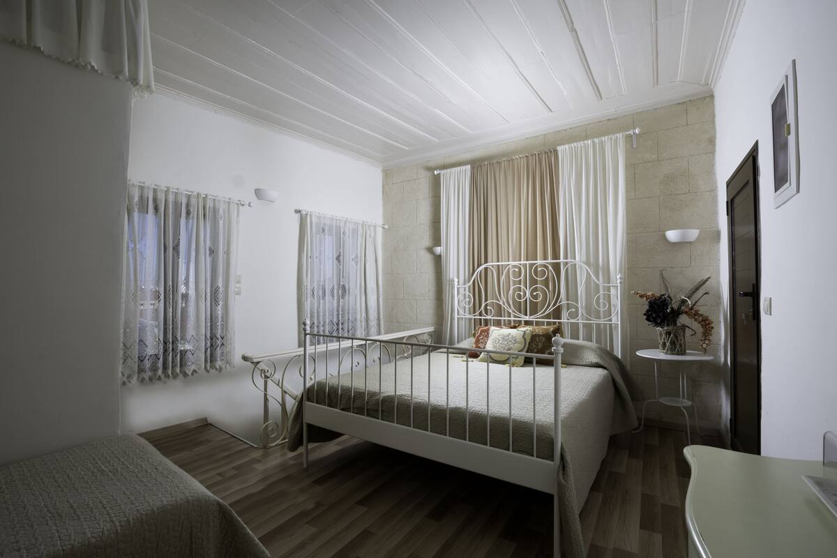 Un dormitorio con cama, una mesita de noche, una pequeña mesa con un jarrón y una ventana con cortinas.