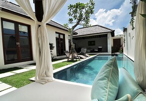 A poolside area with a clear view of a swimming pool, a tree, and a building with large windows and a door. - Arama Riverside (Legian)