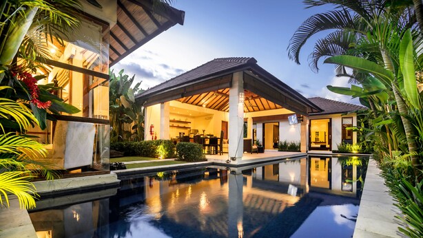 A modern pool area with a covered patio, lush tropical plants, and a clear pool reflecting the surroundings. - Arama Riverside (Legian)