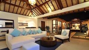 A spacious living area with a high ceiling, a large sofa set, a coffee table, and a dining area with a view. - Arama Riverside (Legian)