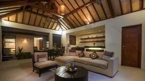 A spacious living area with a high ceiling, a round coffee table, a beige sofa, and a dining area in the background. - Arama Riverside (Legian)